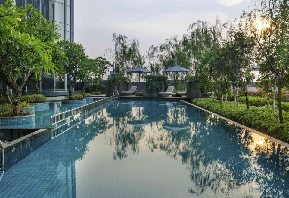 a long , empty swimming pool surrounded by lush greenery , with several lounge chairs placed around the pool at Swissôtel Jakarta Pik Avenue