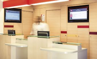 a person in a white lab coat is standing behind a glass counter with two large screens at Ibis Epinay Sur Seine - Gennevilliers