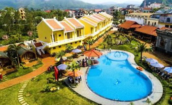 a large yellow building with a swimming pool in front of it , surrounded by green grass and trees at Doan Gia Resort Phong Nha
