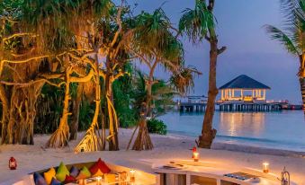 a romantic outdoor dining area with tables , chairs , and cushions is set up on a beach near the water at JW Marriott Maldives Resort & Spa