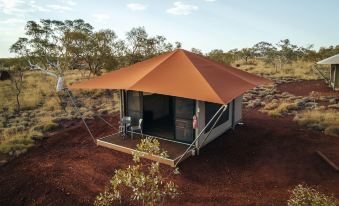 a small , orange tent is set up in a desert - like environment with trees and red soil at Karijini Eco Retreat