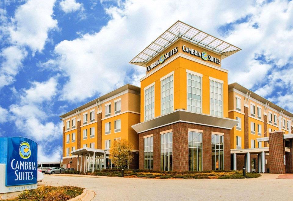 a large hotel building with a blue sign in front of it , surrounded by trees at Cambria Hotel Appleton