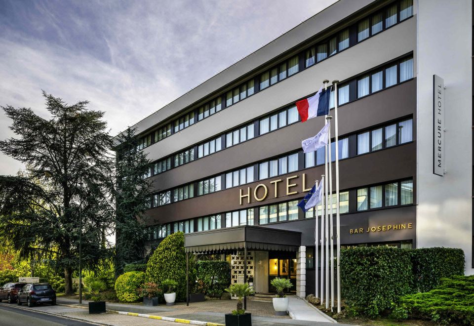 "a modern hotel building with the word "" hotel "" prominently displayed on its side , surrounded by trees and flags" at Mercure Versailles Paris Ouest