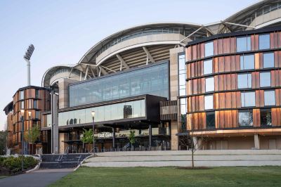 a modern building with a glass facade and a curved roof , surrounded by trees and grass at Oval Hotel at Adelaide Oval, an EVT hotel