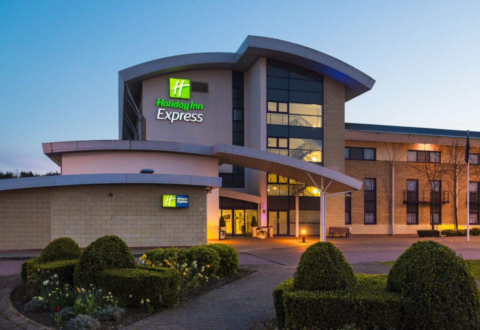 "a modern hotel building with the name "" holiday inn express "" on top , surrounded by greenery and lit up at night" at Holiday Inn Express Northampton - South