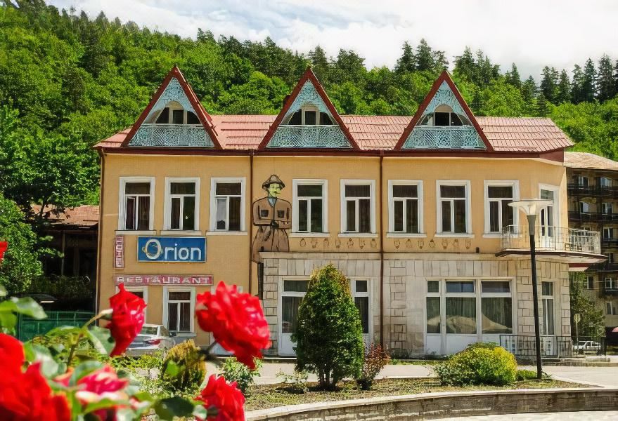 a large yellow building with triangular windows and a red roof is surrounded by flowers at Orion Hotel