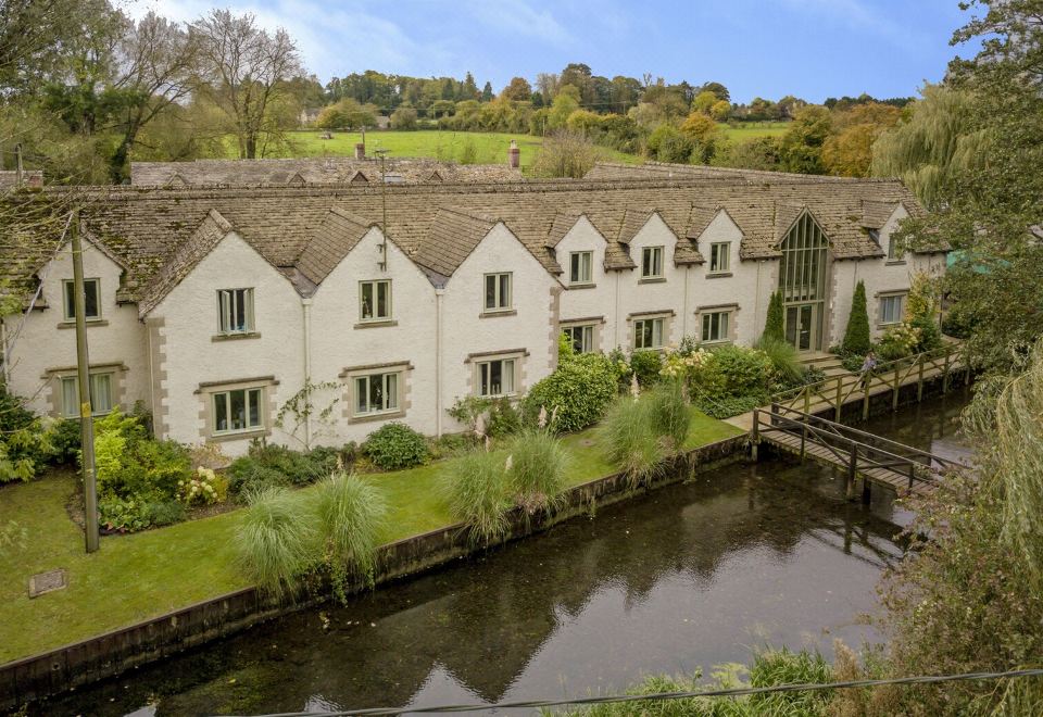 a row of white buildings with brown roofs and green grass surrounding a river , set against a backdrop of trees and hills at Wild Thyme & Honey