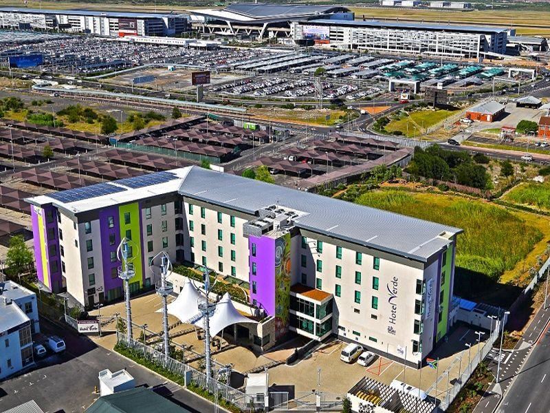 a large , multi - story building with a green and purple facade is surrounded by other buildings and parking lots at Hotel Verde Cape Town Airport