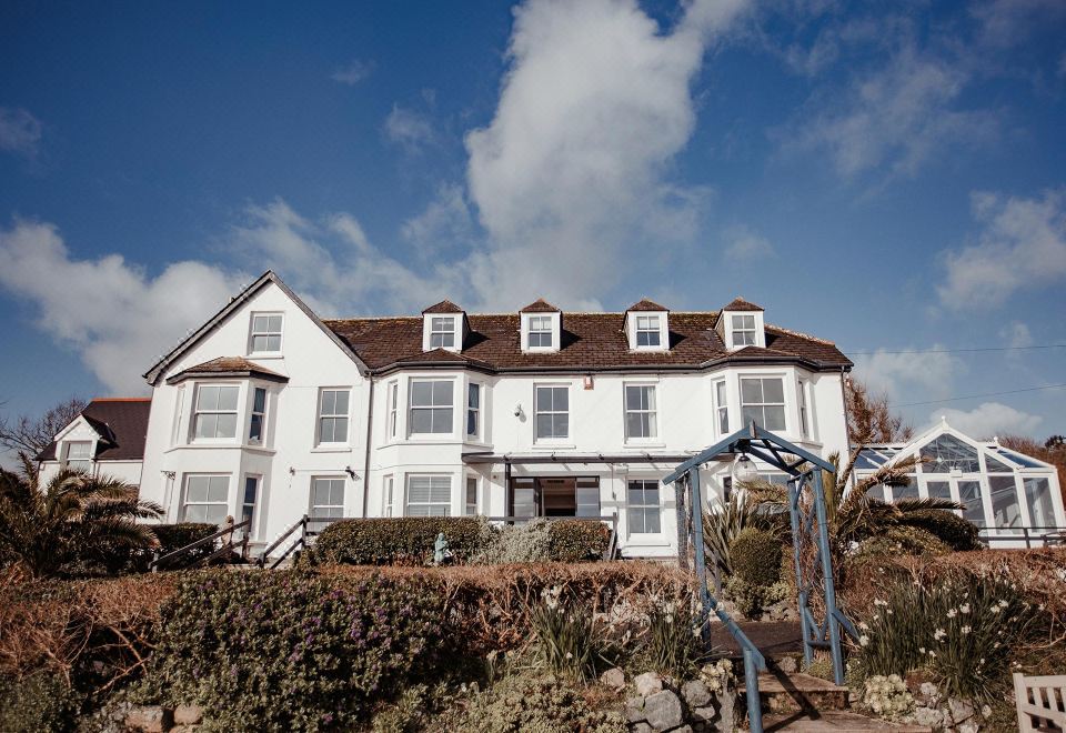 a large white house with a brown roof and bushes in front of it , under a blue sky with clouds at The Bay Hotel