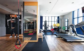 a well - equipped gym with various exercise equipment , including weightlifting machines and mats , under a large window at Garda Hotel