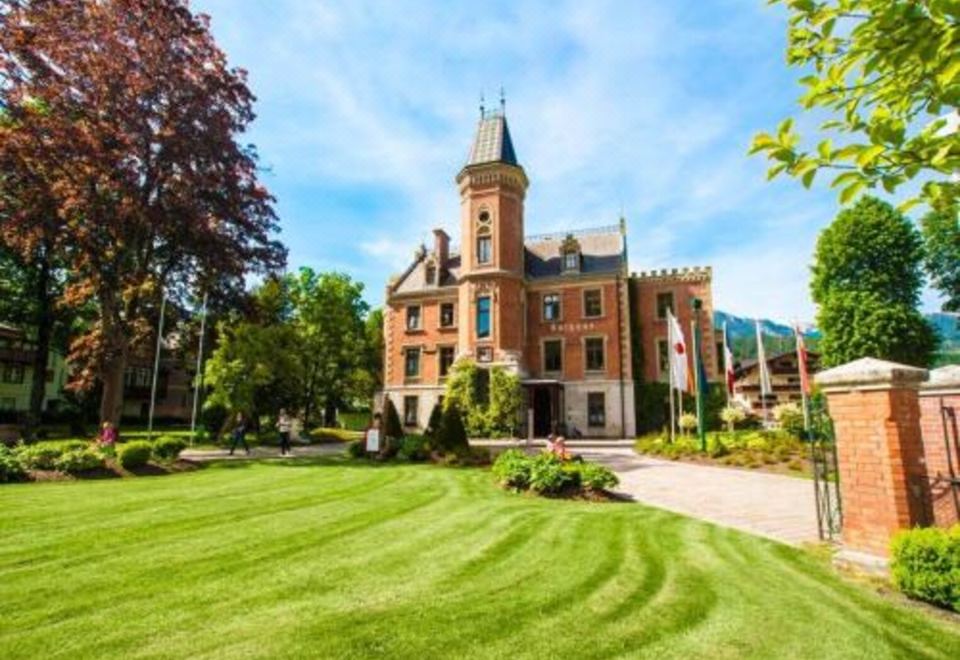 a large red brick building with a clock tower , surrounded by green grass and trees at Hotel Neue Post