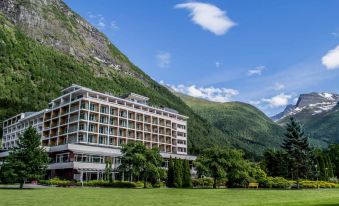 a large , modern hotel surrounded by lush green grass and mountains , with a clear blue sky in the background at Hotel Alexandra Loen