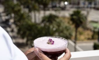 a man in a white shirt is holding a pink drink on a balcony overlooking a cityscape at OKKO HOTELS Toulon Centre