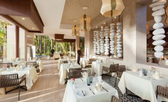 a large dining room with white tables and chairs , all set for a meal , under a thatched roof at Hotel Riu Palace Oasis