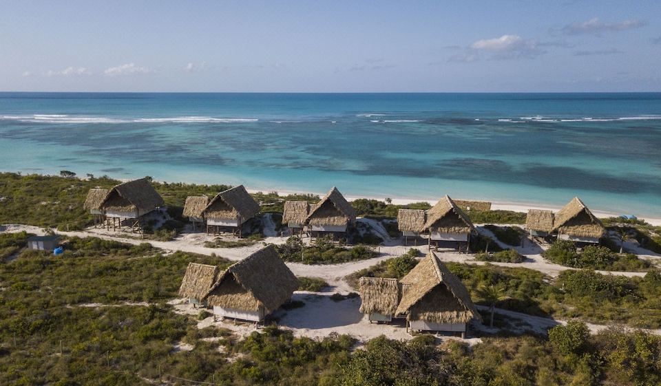 a group of thatched huts on stilts in the ocean , surrounded by sandy beaches and clear blue water at Anegada Beach Club