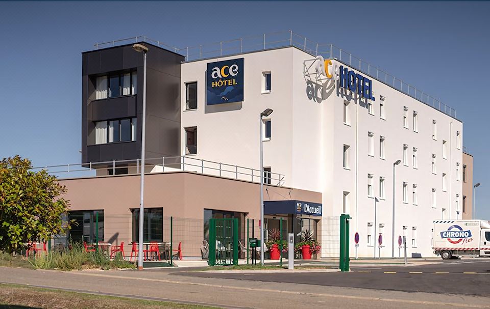 the exterior of the ace hotel , with its sign and surrounding buildings , including tables and green chairs at Ace Hotel le Mans