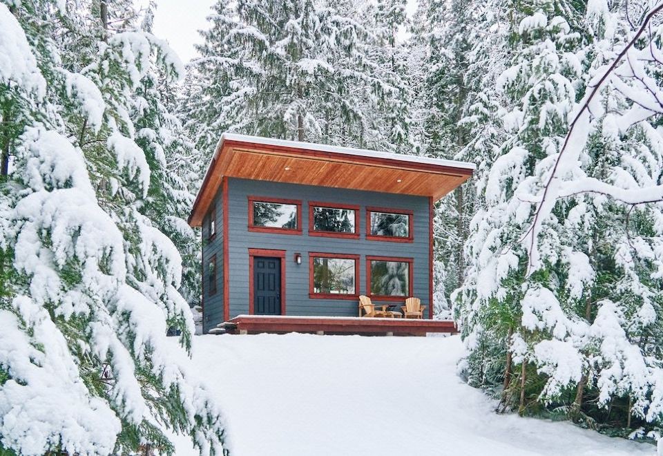 a small wooden cabin nestled in a snowy forest , with a deck and adirondack chairs at Woodland Cabins