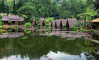 a serene scene of a small pond surrounded by a lush green forest , with several wooden houses dotting the landscape at Imah Seniman