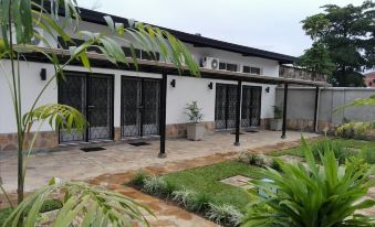 a white building with black shutters , surrounded by green grass and palm trees , under a cloudy sky at Urban Lodge