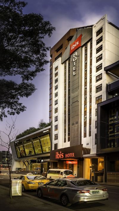 a tall hotel building with a red sign on the front , surrounded by trees and other buildings at Ibis Melbourne Hotel and Apartments