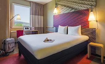 a large bed with white linens and a wooden headboard is in a room with a window , lamp , and nightstand at Ibis Baden Neuenhof