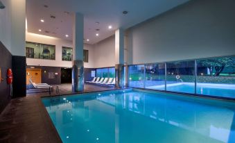 an indoor swimming pool surrounded by lounge chairs , with several people enjoying their time in the pool area at NH Collection Santiago de Compostela