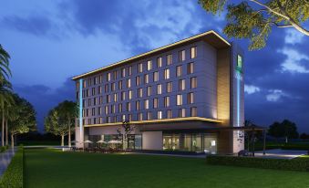 a modern hotel building with a large glass facade , surrounded by trees and grass , at dusk at Holiday Inn Express Kolkata Airport