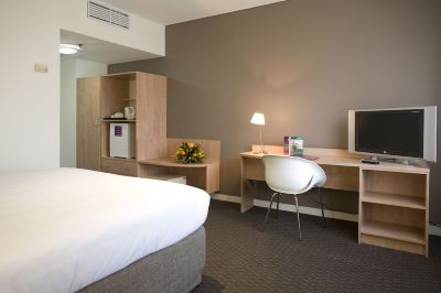 a modern hotel room with a bed , desk , and chair , as well as a lamp and flowers at Ibis Melbourne Hotel and Apartments