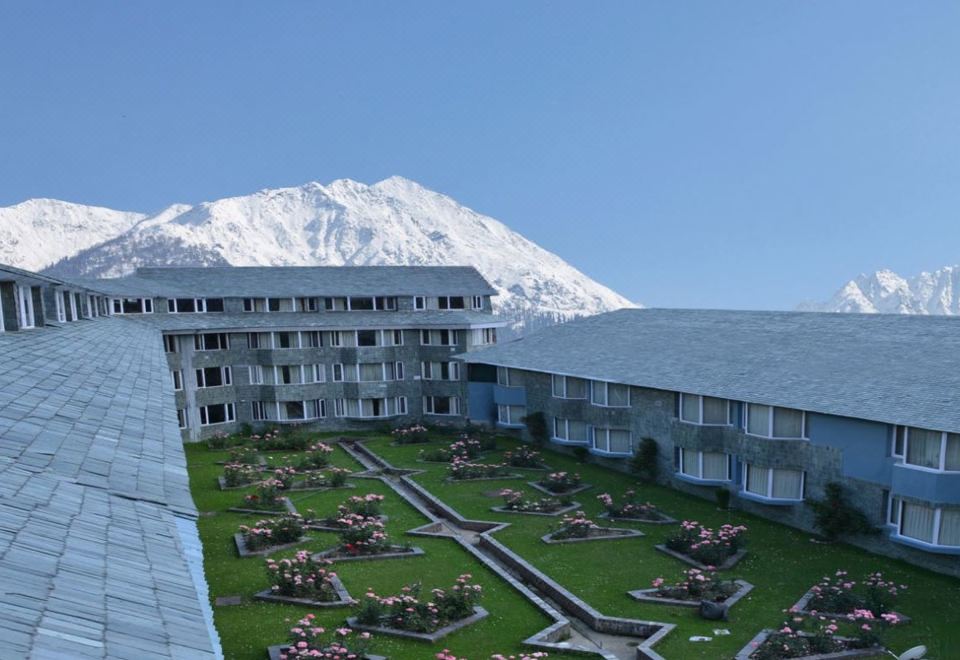 a large building with a green lawn and flower beds is shown in front of a mountain at WoodStock Hotel