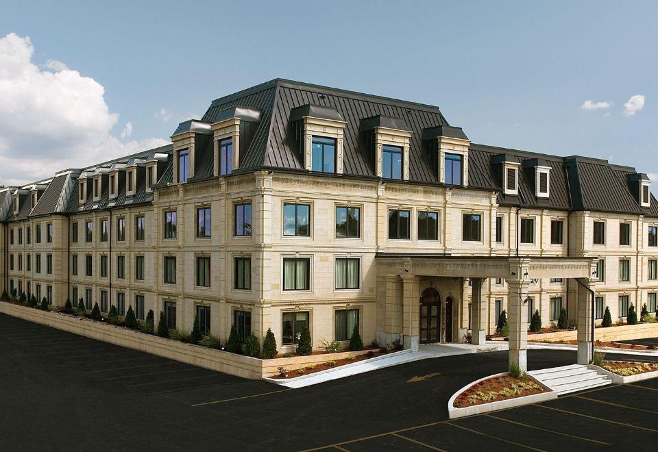 a large , beige building with a curved roof and multiple windows , surrounded by trees and a parking lot at Hotel Brossard