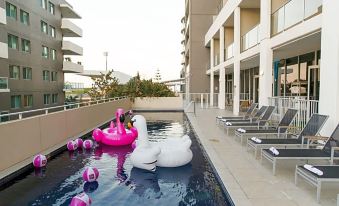 a large swimming pool with a pink flamingo and swan floaties in the middle , surrounded by lounge chairs and a building at Sage Hotel Wollongong