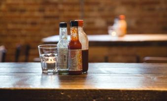 a wooden dining table with two bottles of hot sauce and a small glass on it , along with some bottles in the background at Charter House