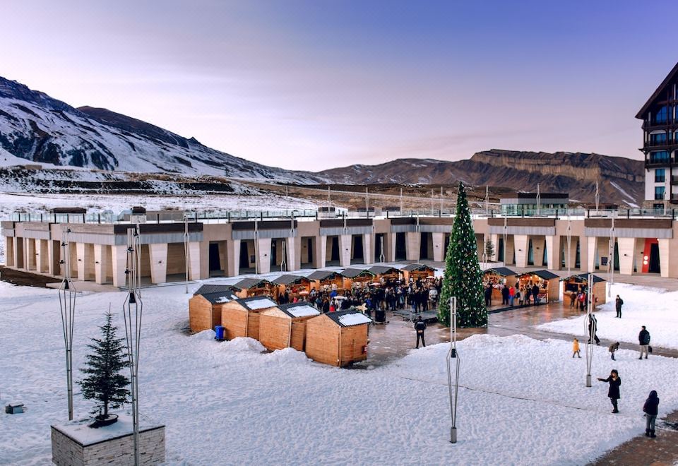 a snowy courtyard with numerous wooden buildings , some of which have trees and people standing around at Shahdag Hotel & Spa