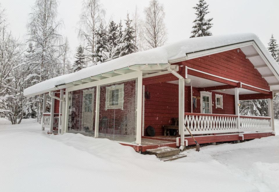 a red wooden house surrounded by snow - covered trees , with a porch and doors visible in the foreground at Kuikka