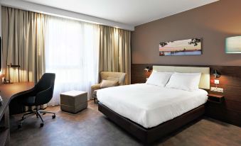 a modern hotel room with a large bed , white linens , and a desk , along with a chair and a window at Hampton by Hilton Toulouse Airport