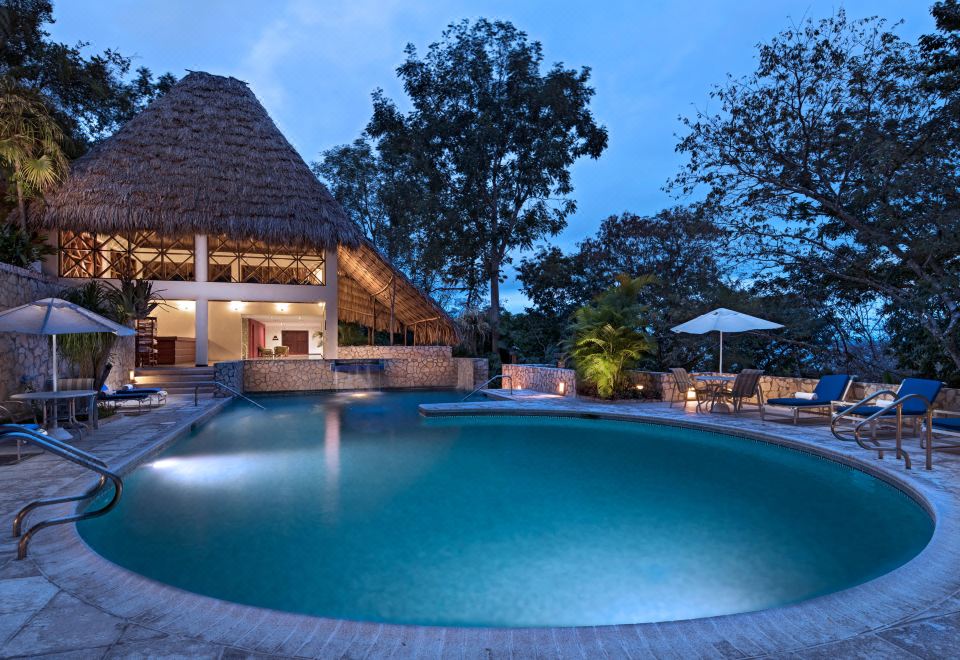 a large outdoor pool is surrounded by a thatched roof structure and trees , with a building in the background at Camino Real Tikal‎