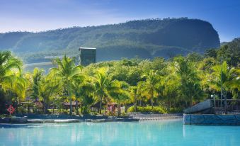 a large swimming pool surrounded by lush greenery and palm trees , with mountains in the background at Rio Quente Resorts - Hotel Pousada