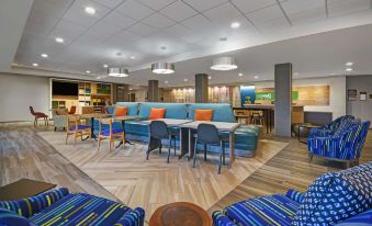 a modern , well - lit hotel lobby with blue and orange couches , wooden floors , and multiple seating areas at Home2 Suites by Hilton Springdale Cincinnati