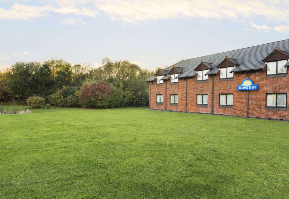 a large grassy field with a brick building in the background , surrounded by trees and bushes at Days Inn by Wyndham Chesterfield Tibshelf