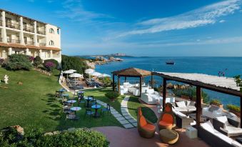 a luxurious resort with a pool surrounded by lush greenery , umbrellas , and outdoor furniture , overlooking the ocean at Club Hotel Baja Sardinia