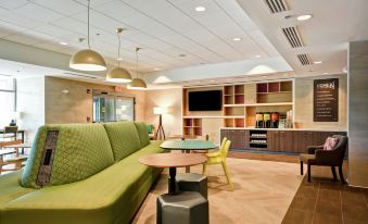 a modern office space with a green couch , wooden desk , and bookshelves , surrounded by large windows and pendant lights at Home2 Suites by Hilton Walpole Foxboro