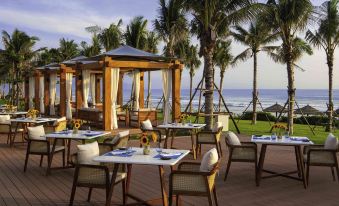 an outdoor dining area with several tables and chairs , surrounded by palm trees and overlooking the ocean at Movenpick Resort Cam Ranh