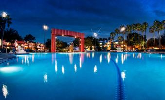 a large blue swimming pool illuminated at night , with a red archway in the background at Disney's All-Star Movies Resort