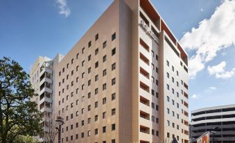 a modern , beige - colored building with multiple floors and balconies , surrounded by trees and other buildings under a clear blue sky at Mitsui Garden Hotel Okayama