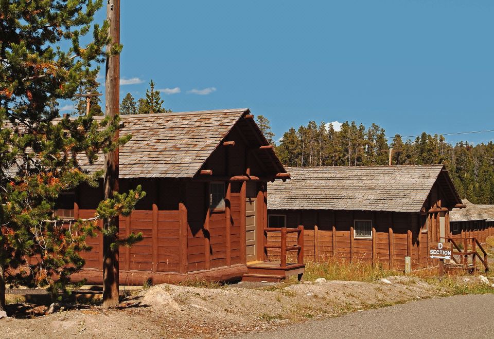 a wooden cabin is situated next to a stone wall and surrounded by trees , with a dirt road leading up to it at Lake Lodge