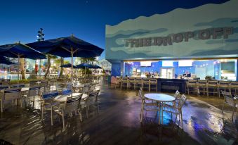 "a nighttime view of a restaurant with tables and chairs , an umbrella , and the word "" the shop "" on the building" at Disney's Art of Animation Resort