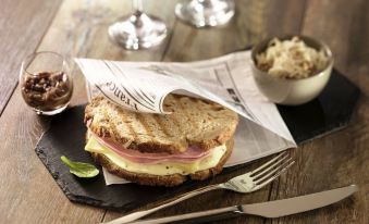 a sandwich with cheese and ham is on a slate tray , accompanied by a fork , knife , and wine glass at Ibis Epinay Sur Seine - Gennevilliers