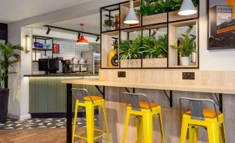 a modern café with wooden tables and yellow stools , green plants , and hanging lights , as well as a coffee bar area at Super 8 by Wyndham Chester East