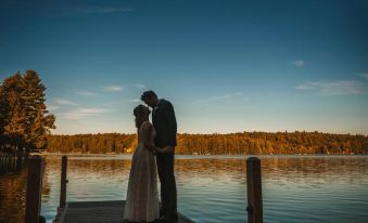 a newlywed couple standing on a wooden dock , embracing and kissing , with the sun setting in the background at The Fern Lodge