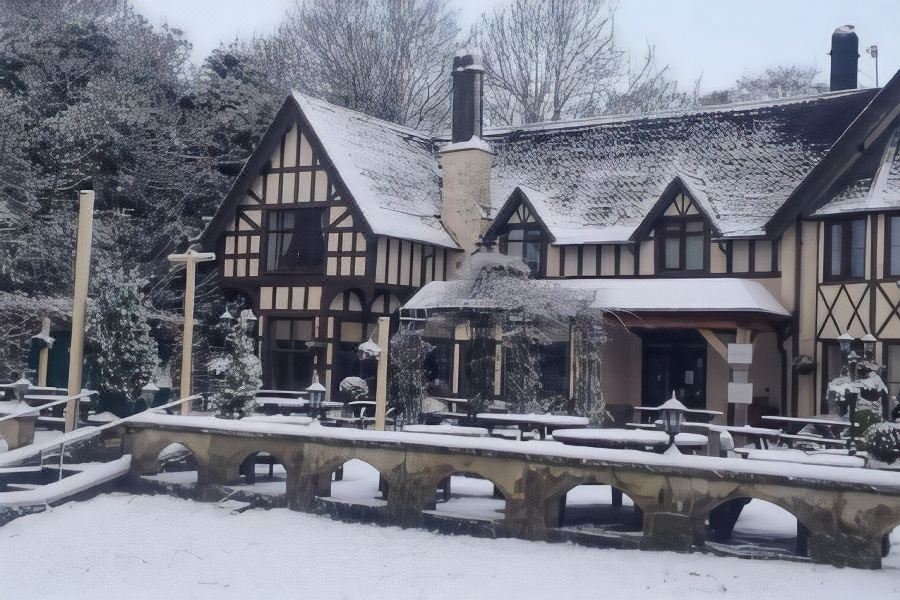 a snow - covered house with a wooden walkway and benches in front of it , surrounded by trees at The Bentley Brook Inn